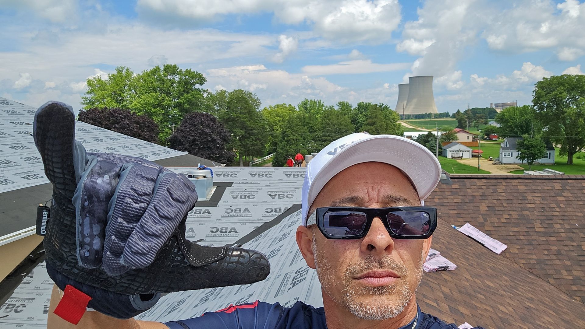 Man taking a selfie on a rooftop with a stone city sign, wearing sunglasses and a white cap.