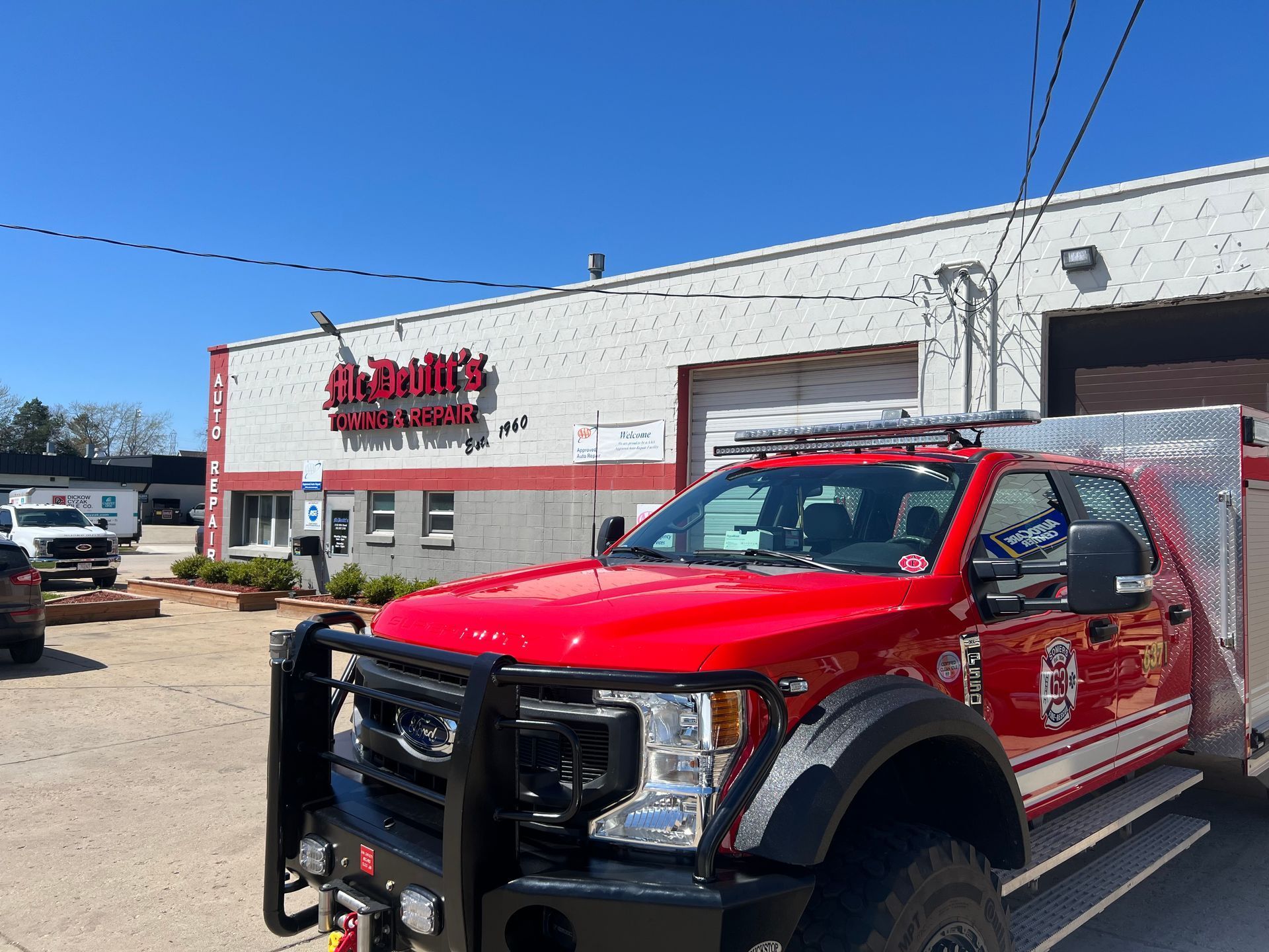 A bright red Ford fire department truck parked in front of a white warehouse building under a clear blue sky.