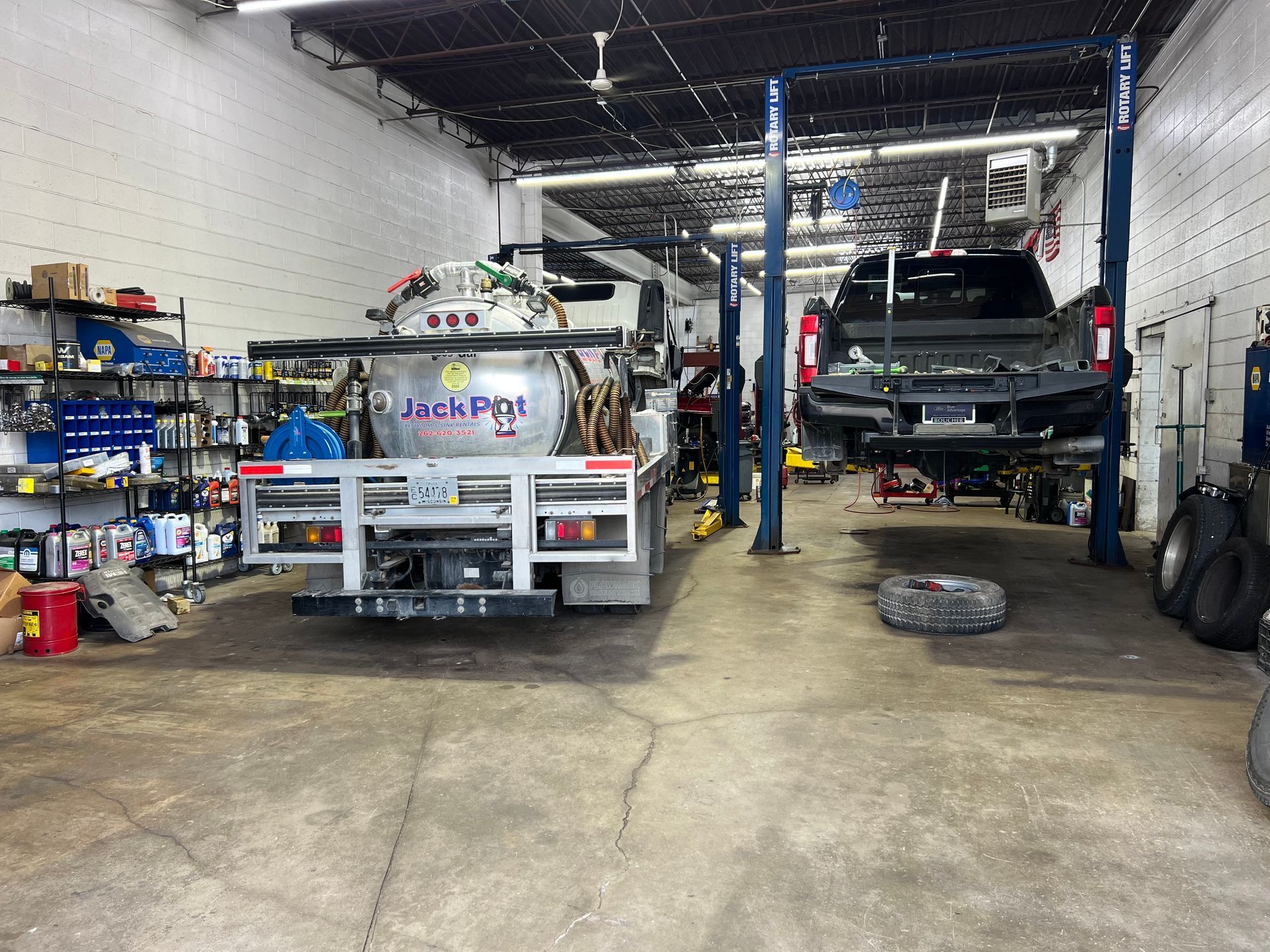 A busy vehicle repair shop with a large industrial tank truck parked on the left and a truck raised on a hydraulic lift.