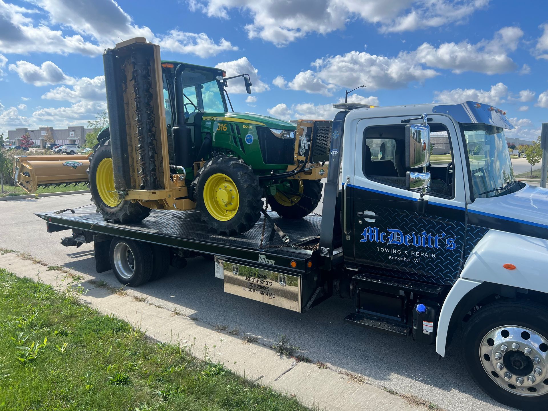 A green John Deere tractor with a side-mounted mower loaded on the flatbed of a tow truck parked on the roadside.