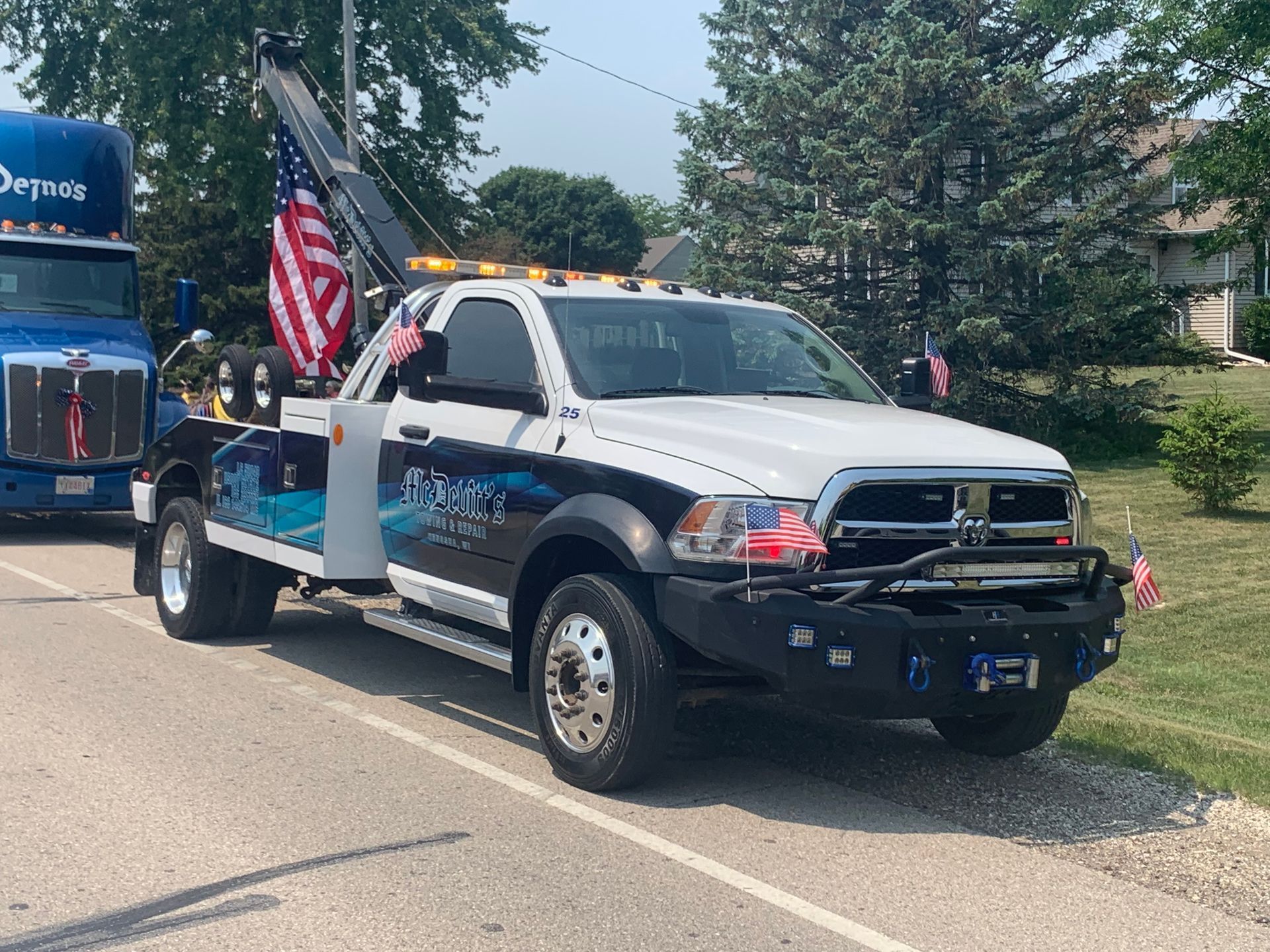 A white Ram tow truck with patriotic American flag decals parked on a roadside next to a blue semi-truck.