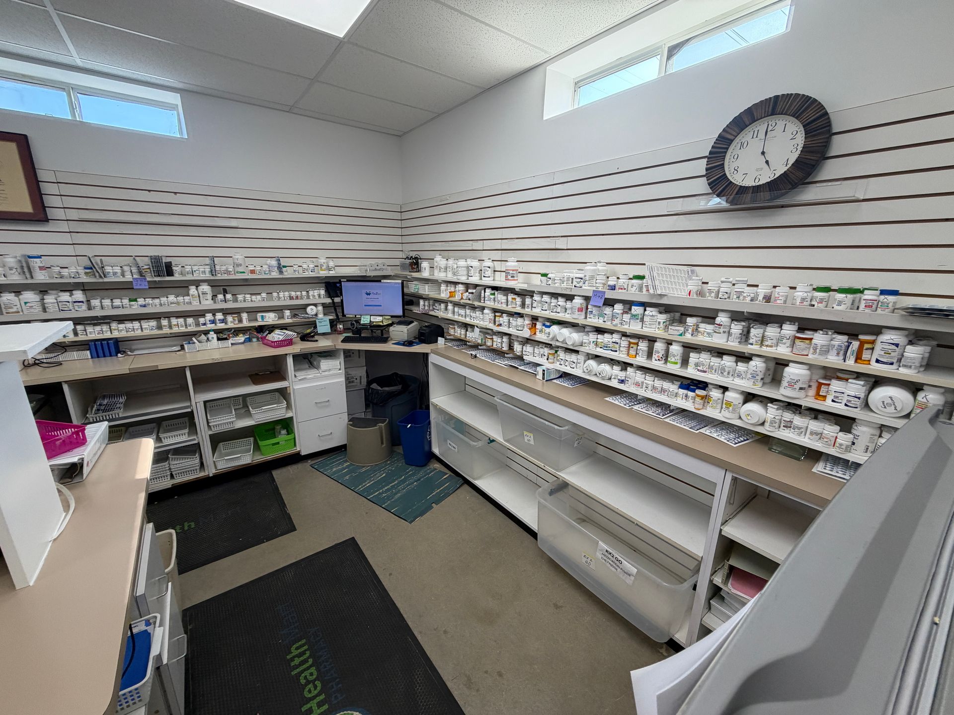 Small retail pharmacy with white shelves of medicines, a computer station, and a wall clock.