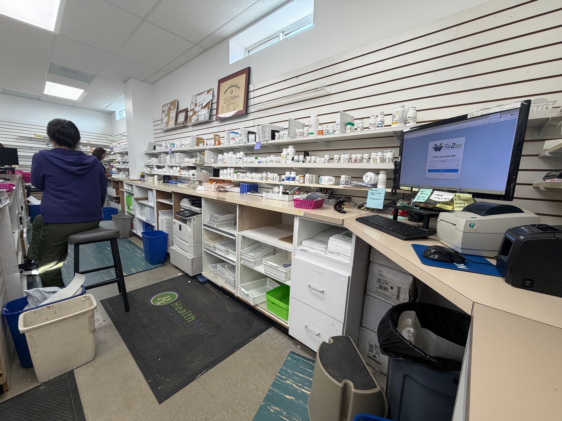 Small pharmacy with shelves of medications, a computer monitor, and a person working at the counter.