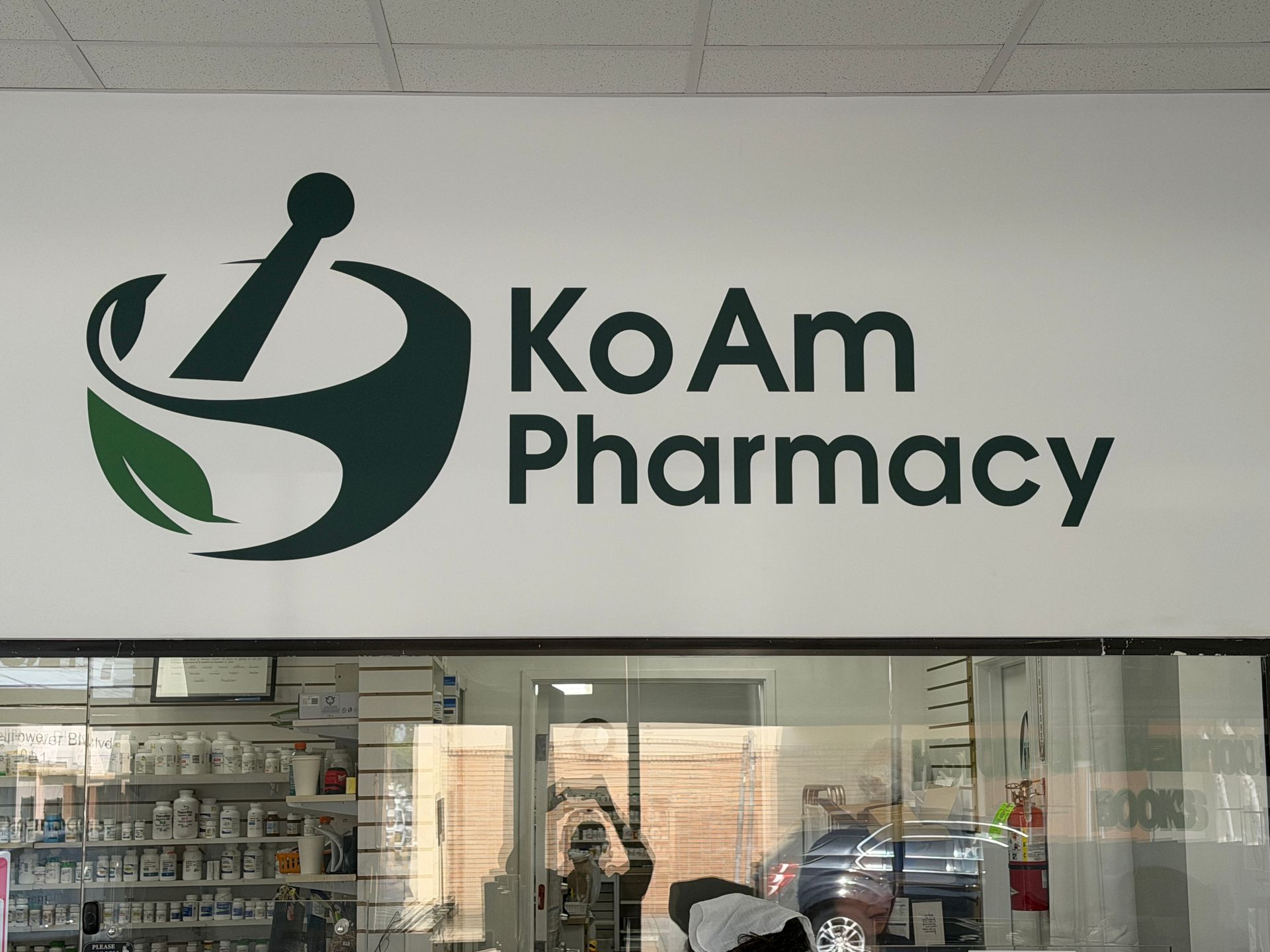 KoAm Pharmacy storefront sign with green logo above glass doors in a mall-like corridor