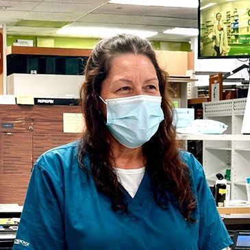 Masked worker in blue scrubs standing in a store stockroom, smiling at the camera.