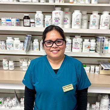 Smiling pharmacy worker in blue scrubs standing in front of shelves of medication bottles