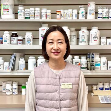 Woman in a lavender vest standing in a pharmacy aisle with shelves of medicine bottles behind her