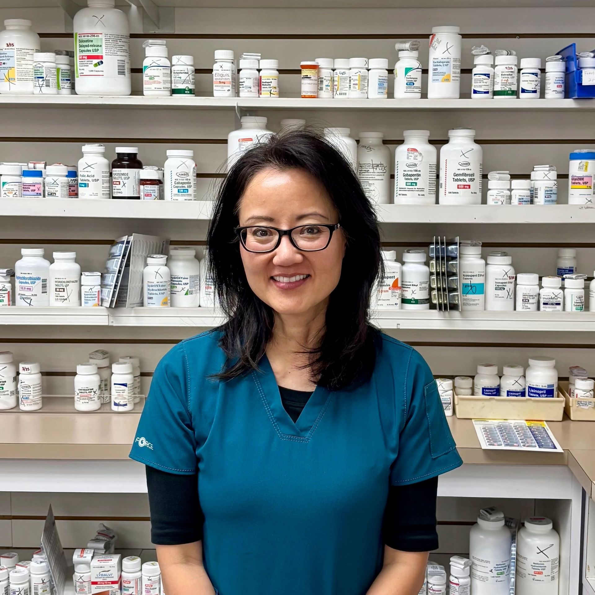 Woman in teal scrubs smiles in a pharmacy, standing before shelves of medicine bottles.