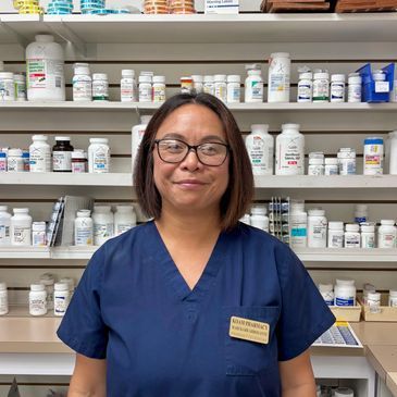 Pharmacist in navy scrubs standing in a pharmacy with shelves of medicine bottles behind her