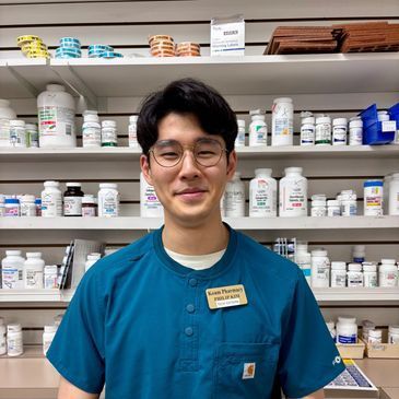 Smiling pharmacy worker in teal scrubs standing in front of stocked medication shelves