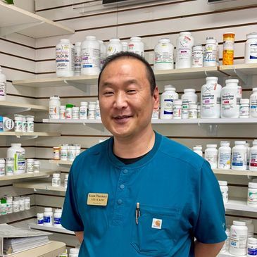 Person in blue scrubs standing in a pharmacy aisle with shelves of medicine bottles.