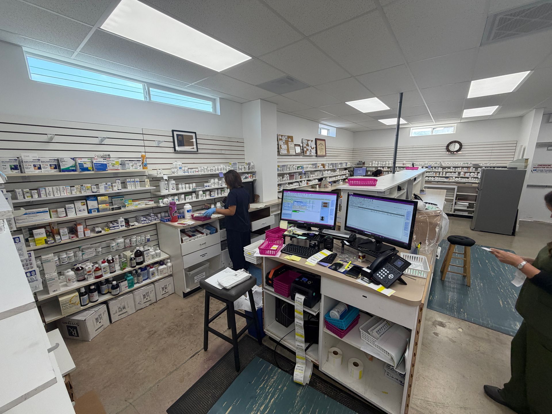 Convenience store checkout counter with monitors, shelves of goods, and a clerk behind the register.
