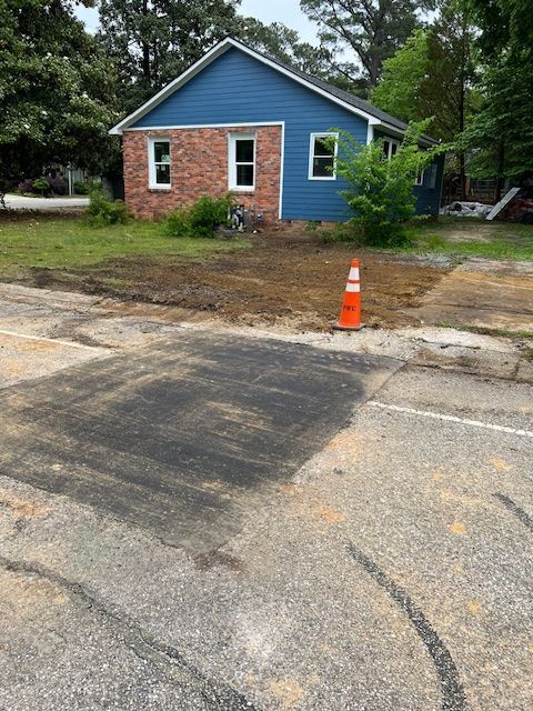 A small house with blue siding and a brick section sits beside a rough parking area with an orange cone.