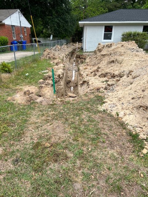 Trench dug in a grassy yard, with visible white pipes; dirt pile and small house in background.