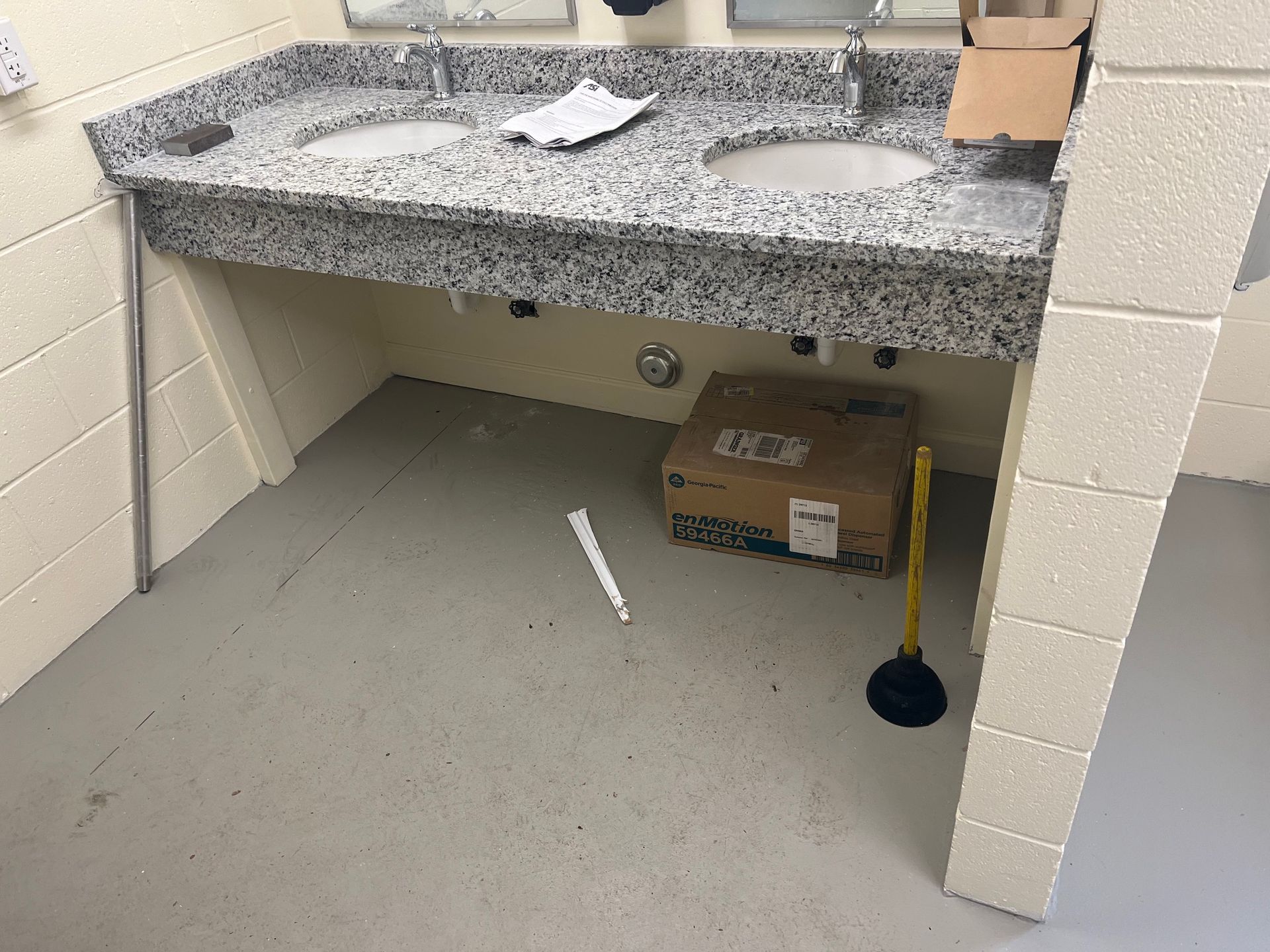 Public restroom with granite countertop, two sinks, and a box under the sinks. A plunger is on the floor.