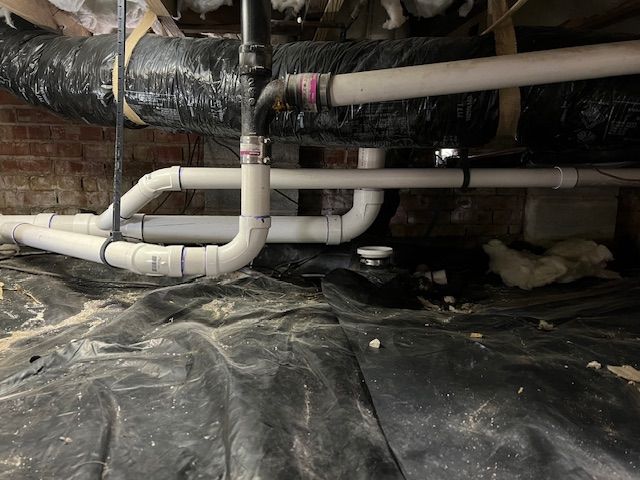 Pipes under a house with black insulation and a black ground cover. Brick wall visible in background.