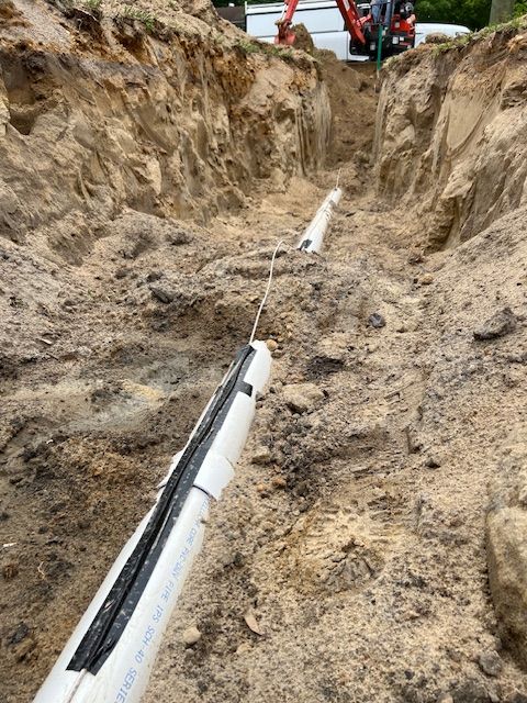 Trench dug in dirt, containing white PVC pipes, a cable, and an excavator in the distance.