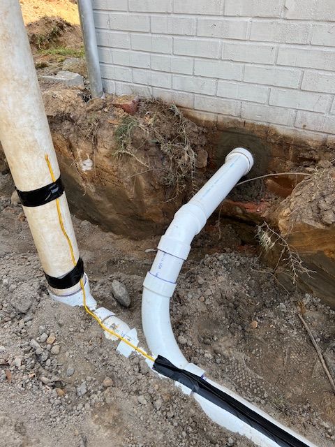White PVC pipes and a beige pipe in a trench near a brick wall, some pipes are taped.