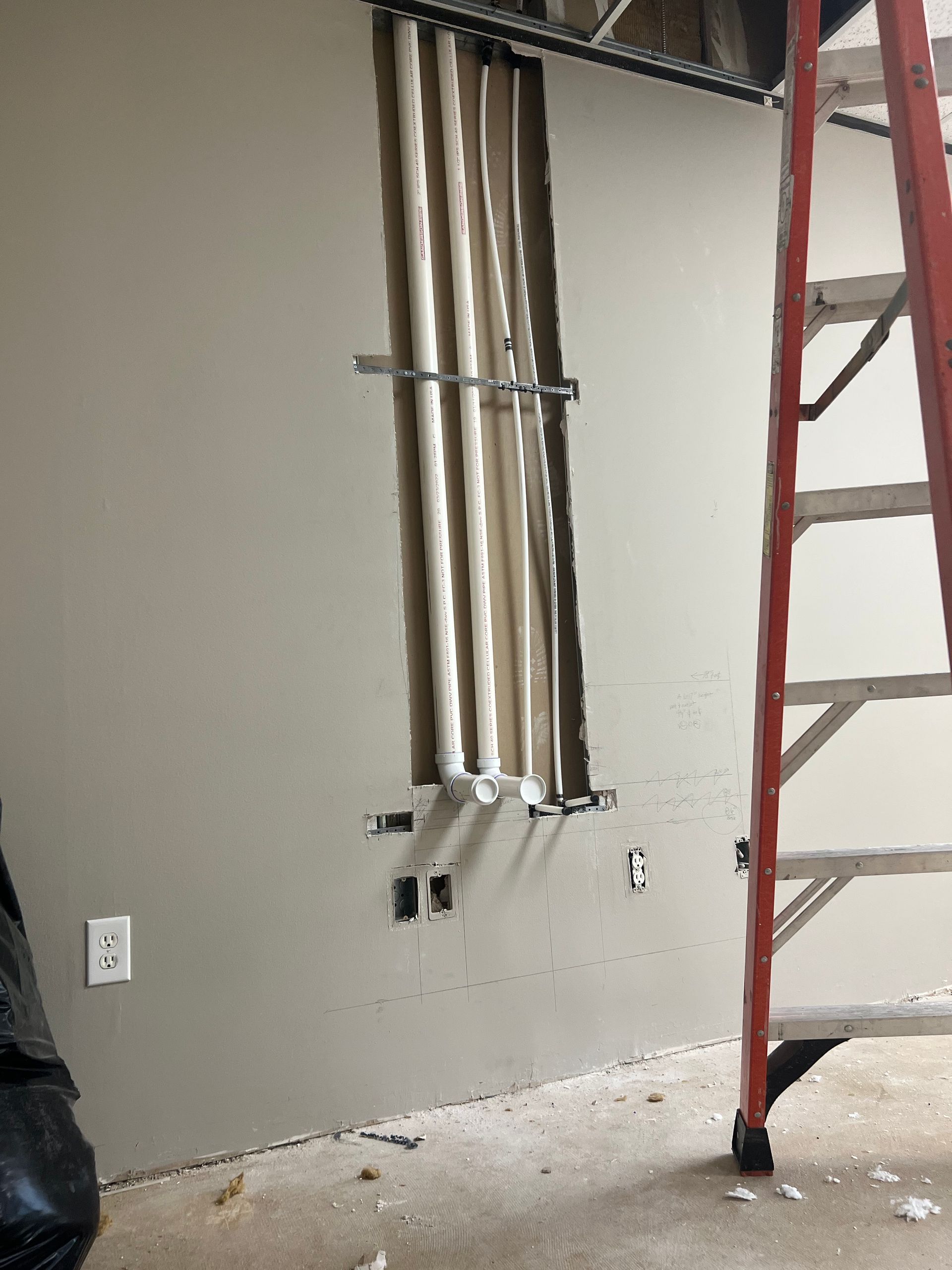 Wall with an exposed section revealing pipes, near a red ladder, and an electrical outlet. Construction in progress.