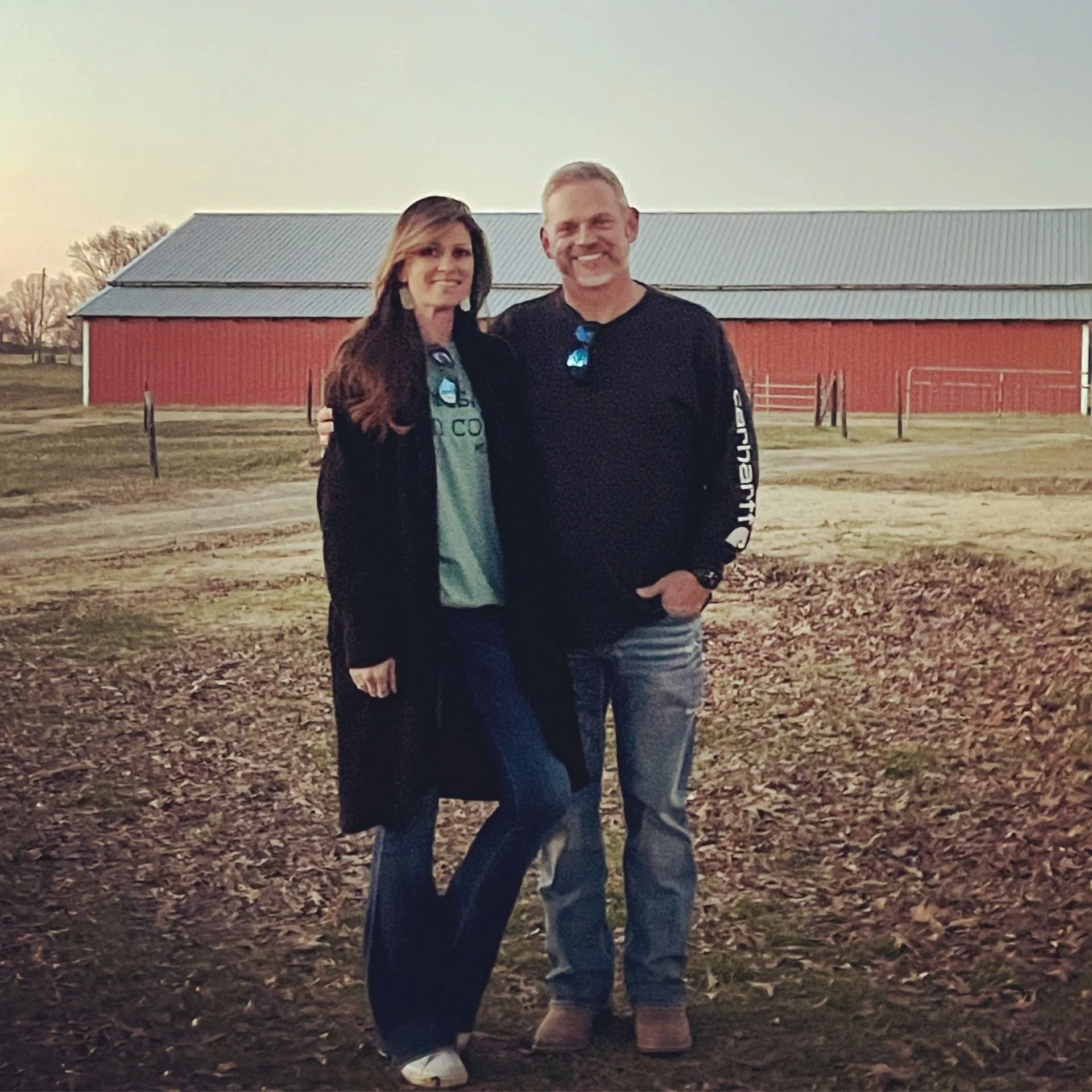 Couple standing in front of a red barn. Woman in coat, jeans; man in jeans, long-sleeved shirt. Outdoors.