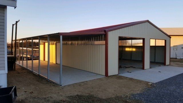 A metal shed with a red roof, two garage doors, and a covered side area, all on a concrete slab.