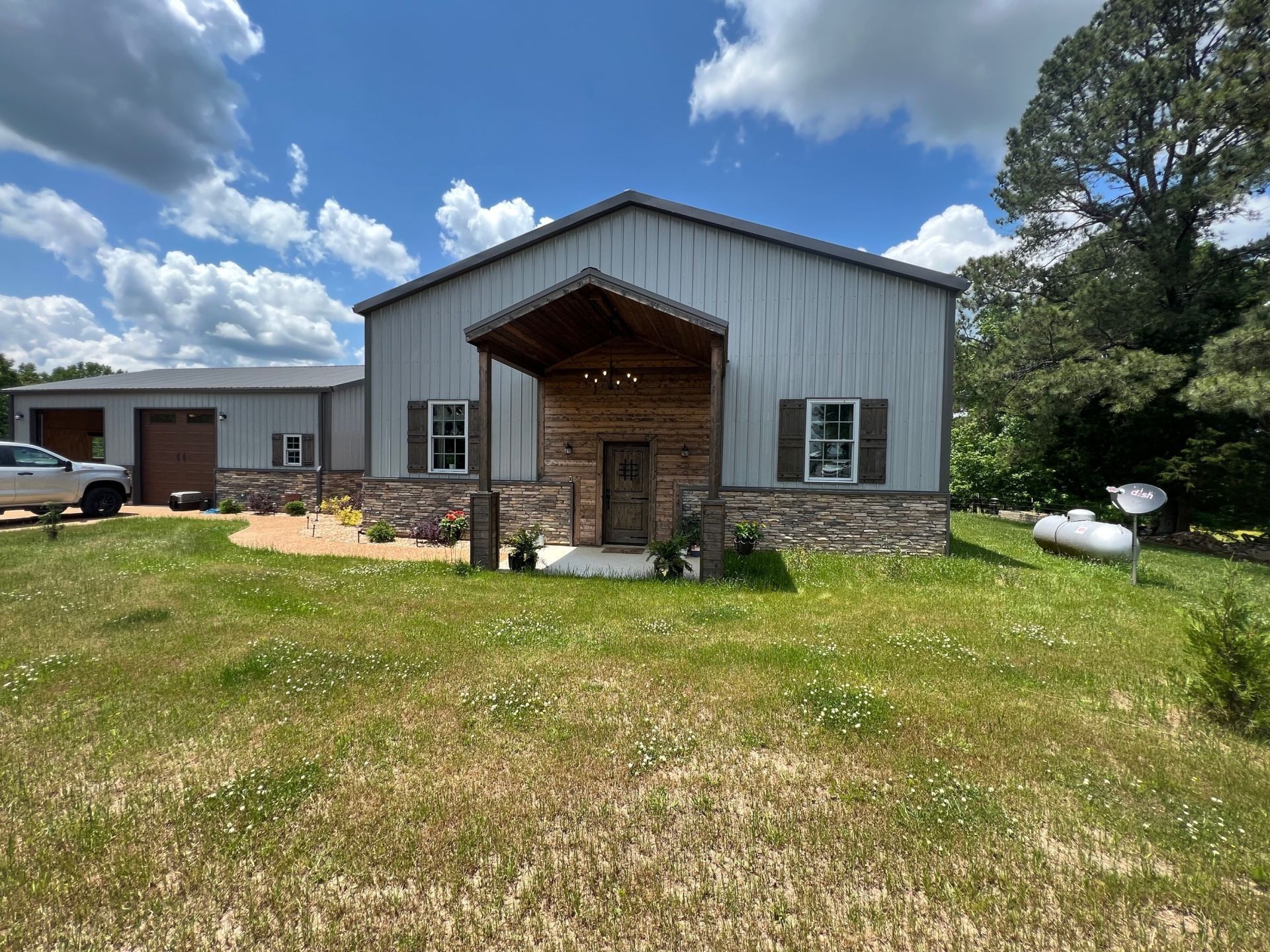 Metal building with a stone facade, porch, and attached garage on a grassy lot under a cloudy sky.