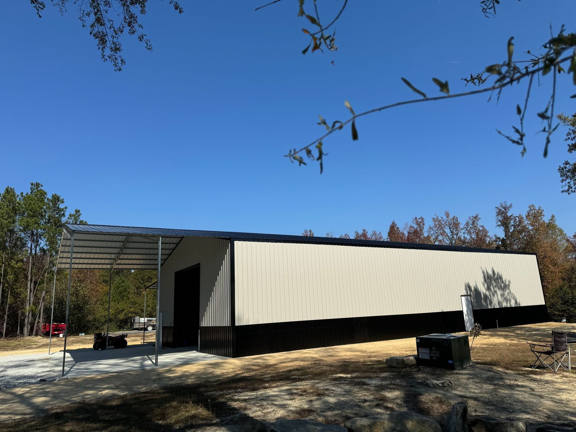 Large, light-colored metal building with a dark roof and a carport on a gravel lot under a blue sky.