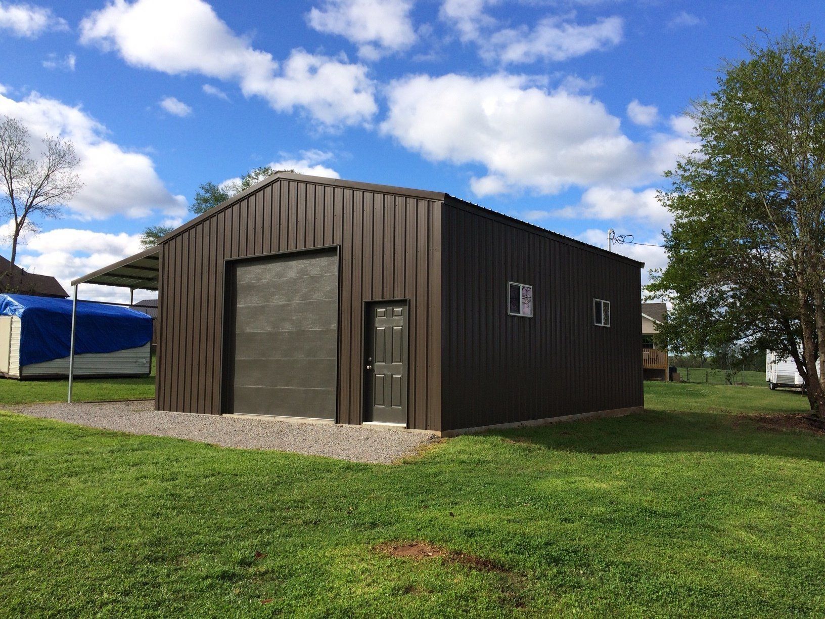 Brown metal garage with large door and small door, two windows, on a grassy plot under a blue sky.
