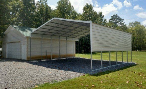 Metal carport with a gravel base attached to a small white building with a green roof.