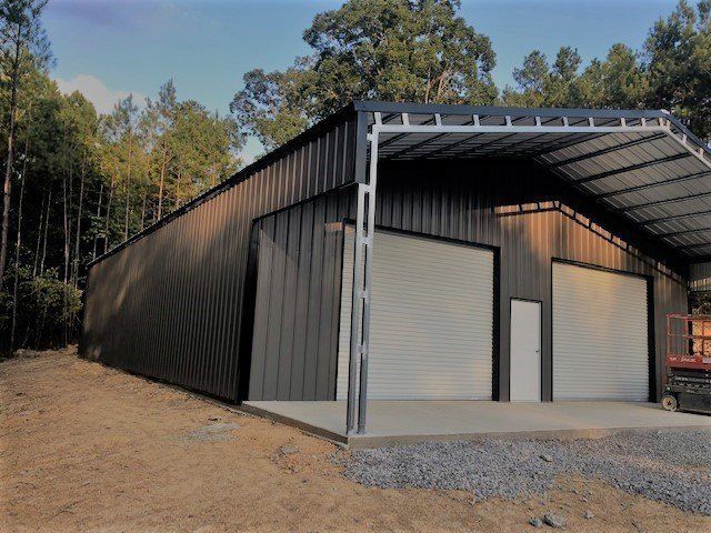 Dark metal building with two garage doors and an attached carport, set in a wooded area.
