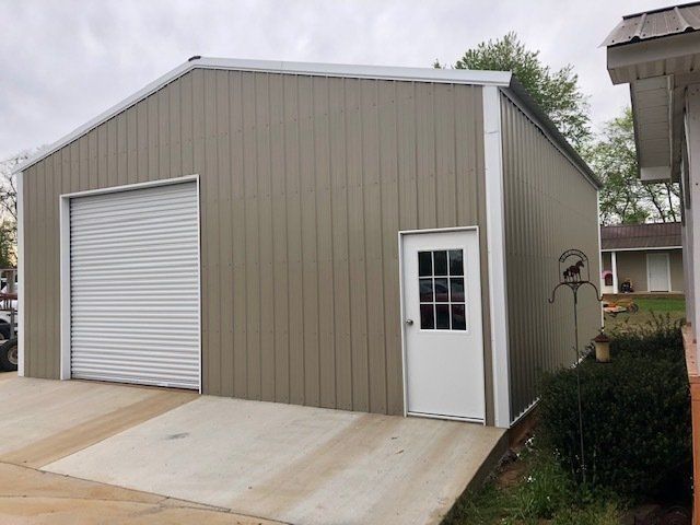 Tan metal building with a garage door and white door. Concrete driveway slopes upward.
