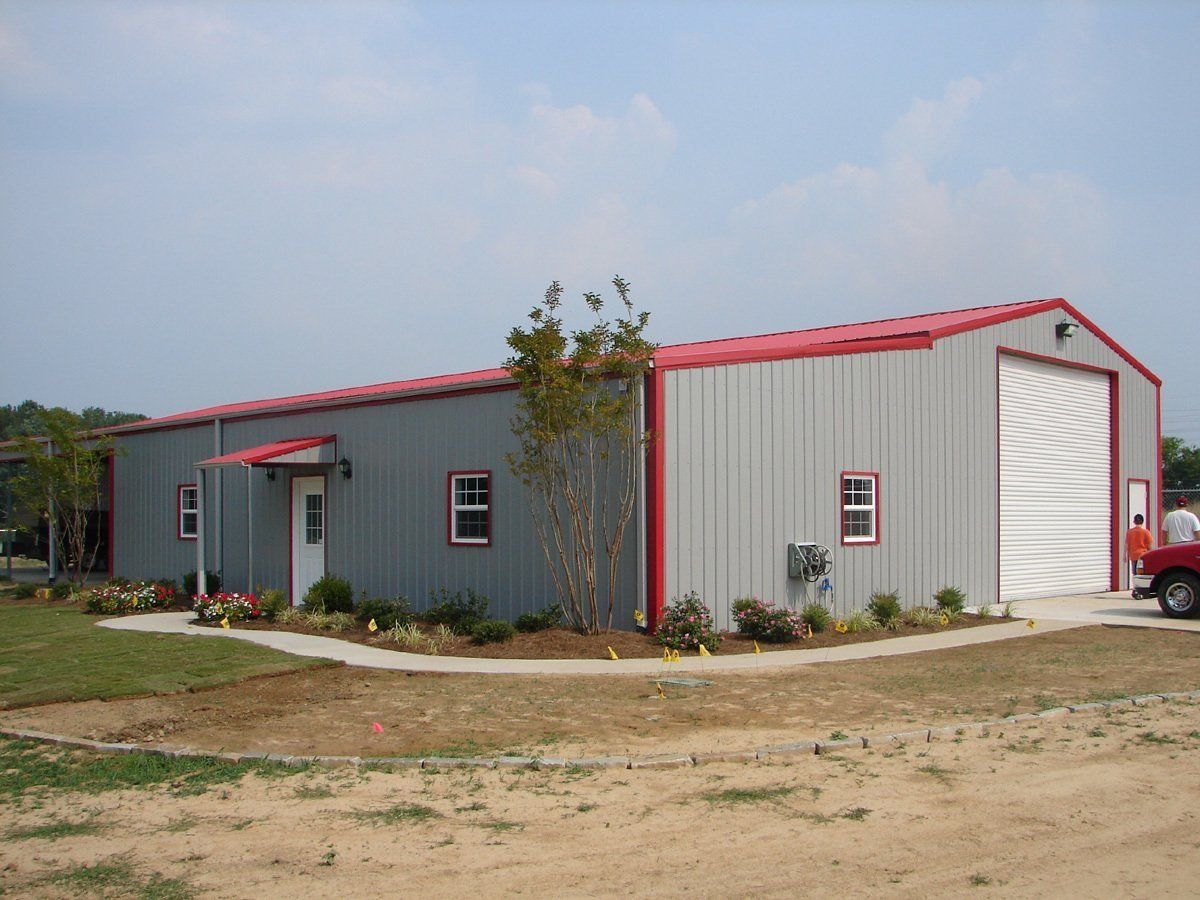 Gray metal building with red trim, roll-up door, small windows, and landscaping.
