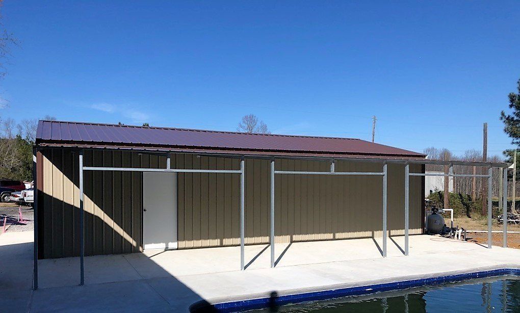 A metal shed with a brown roof sits by a pool. Metal supports hold up an awning. Blue sky.