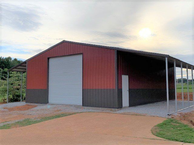 Red and brown metal barn with a roll-up door, a side door, and a covered awning, gravel driveway.