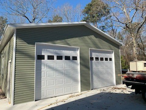 Green-sided garage with two white garage doors, set against a backdrop of bare trees and a blue sky.