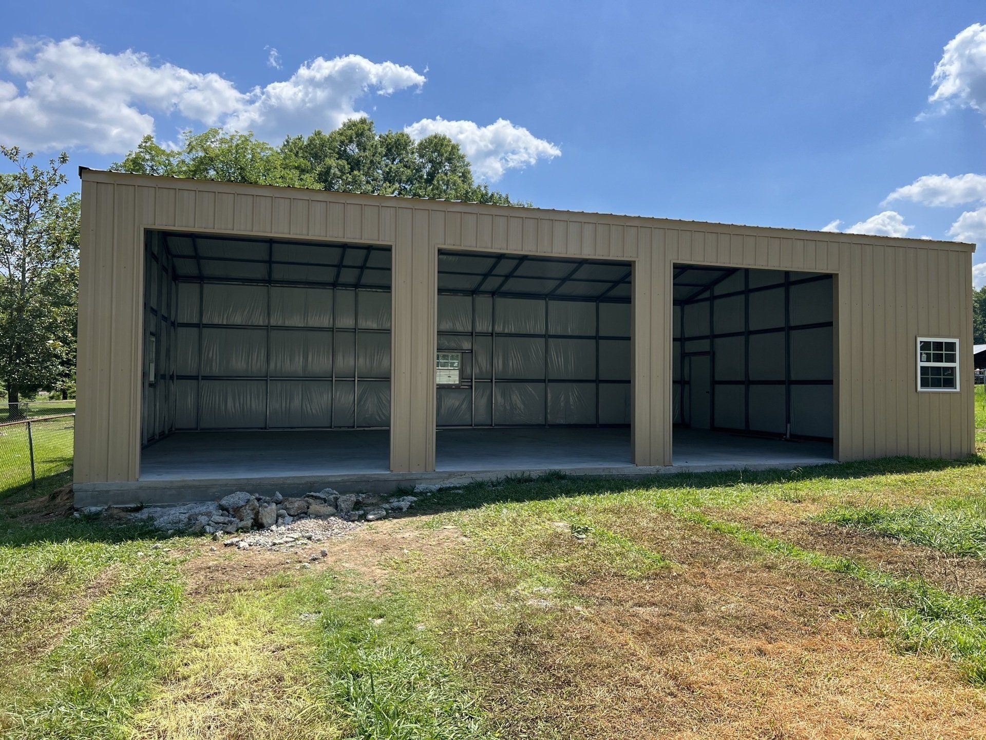 Tan metal carport with three bays, on a concrete slab, set in grassy area under a blue sky.