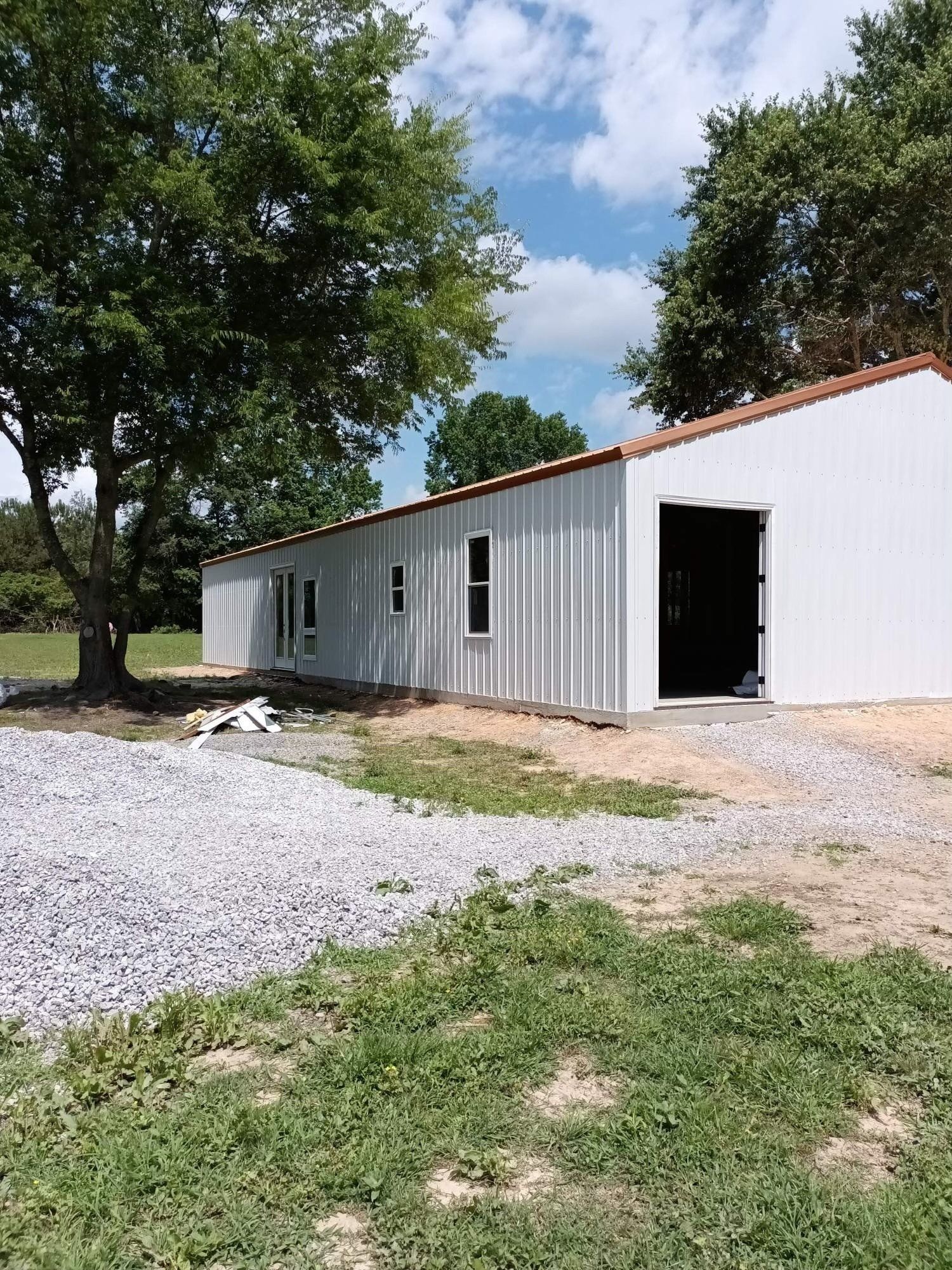 White metal building with open garage door, windows, gravel driveway, and trees.