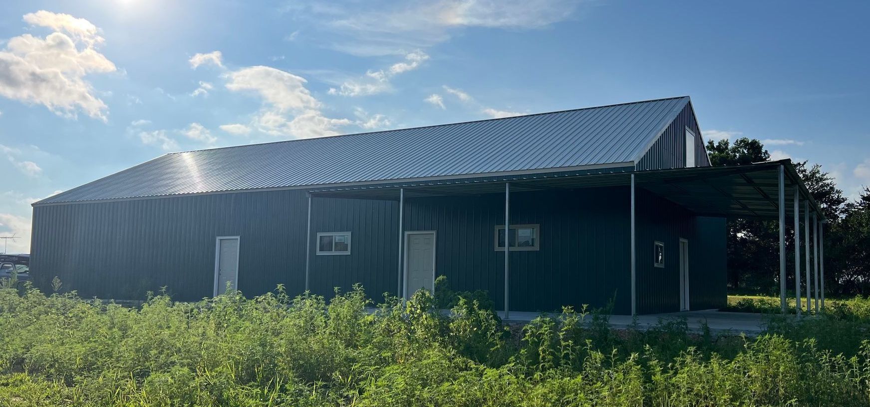 Dark green metal building with a slanted roof and a carport, set in a field under a bright blue sky.