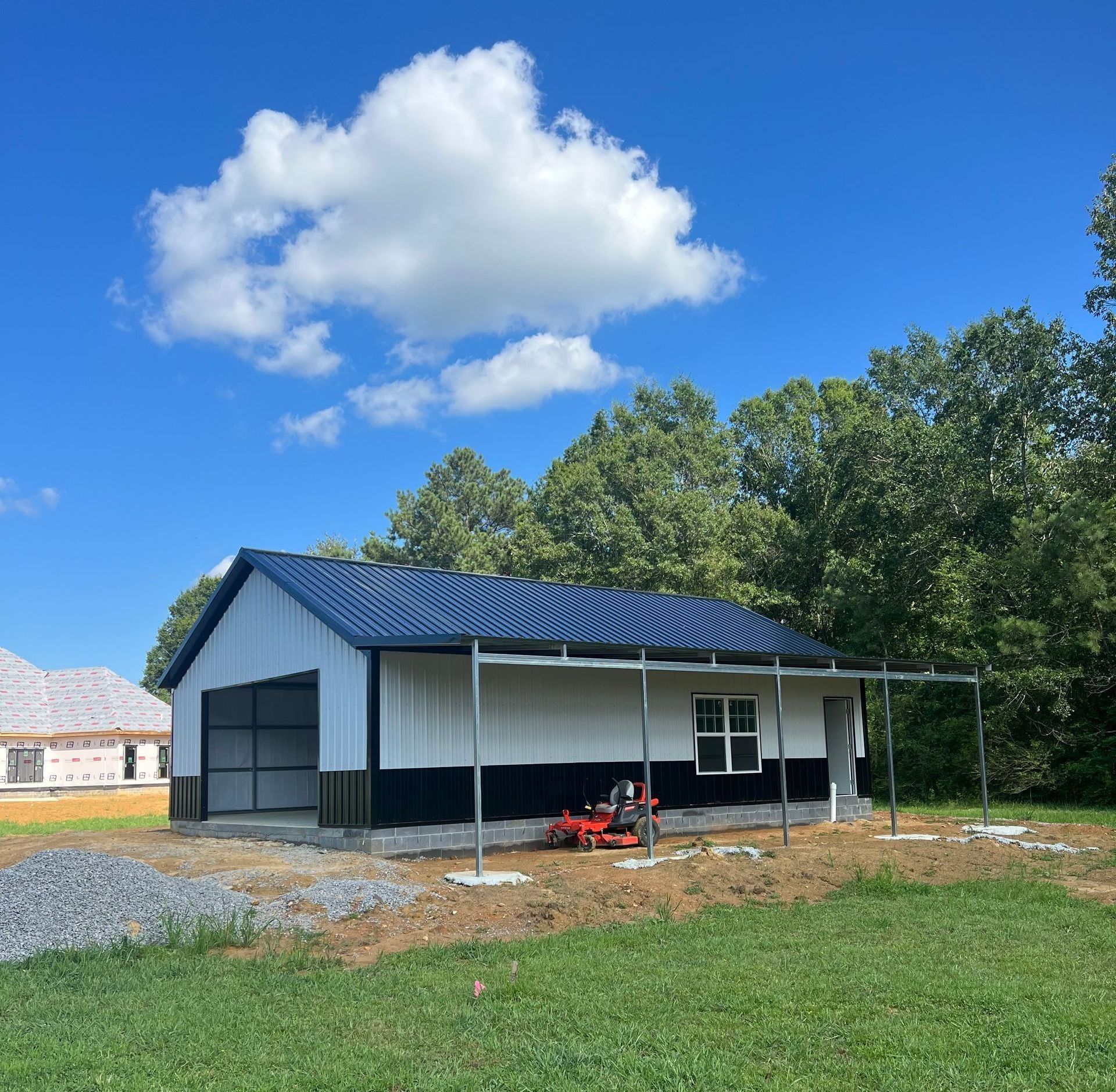 Metal-clad building with black and white siding, blue roof. Small porch with a lawnmower, in front of trees and a blue sky.