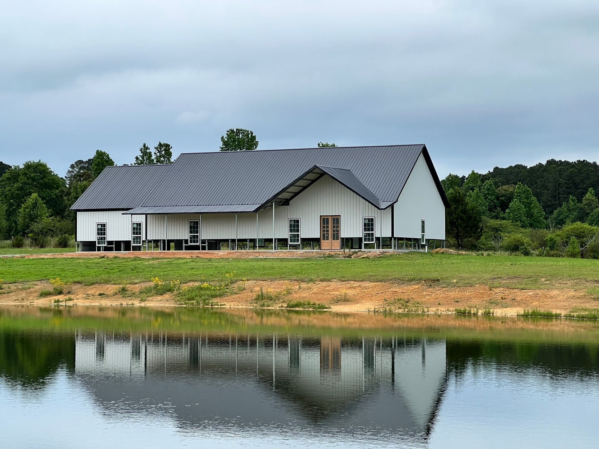 White barn-like building with dark roof reflecting in a calm body of water; set in a green field under a cloudy sky.