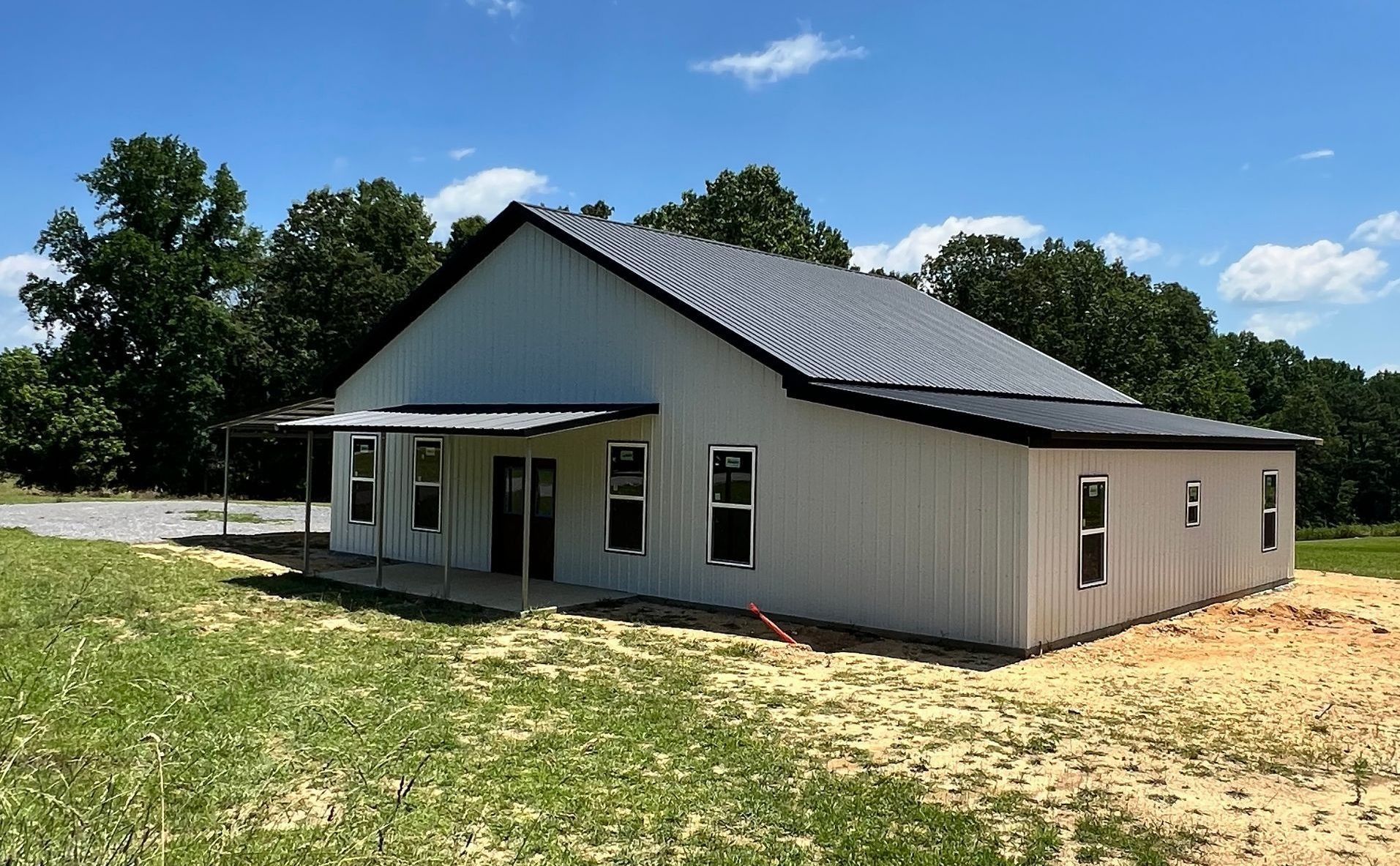 A light gray metal building with a black roof and porch, set in a grassy field with trees.
