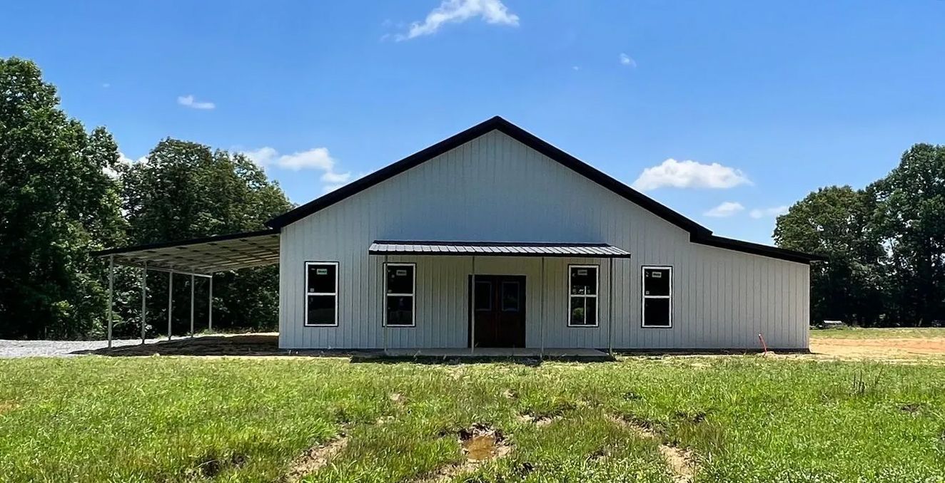 White metal barn with black trim, windows, and a porch, set in a grassy field under a blue sky.