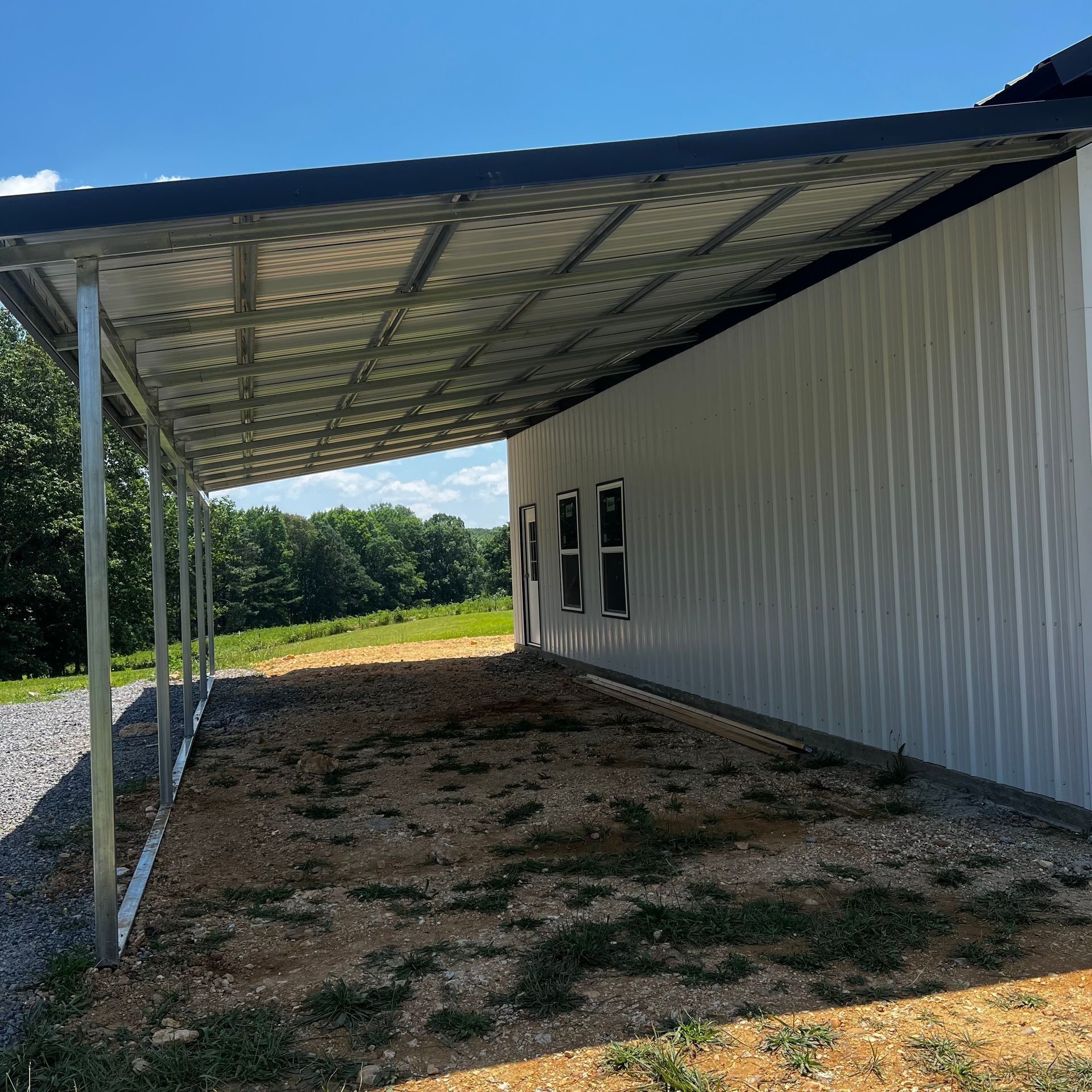 Metal carport attached to a white building with windows, set on gravel and dirt.