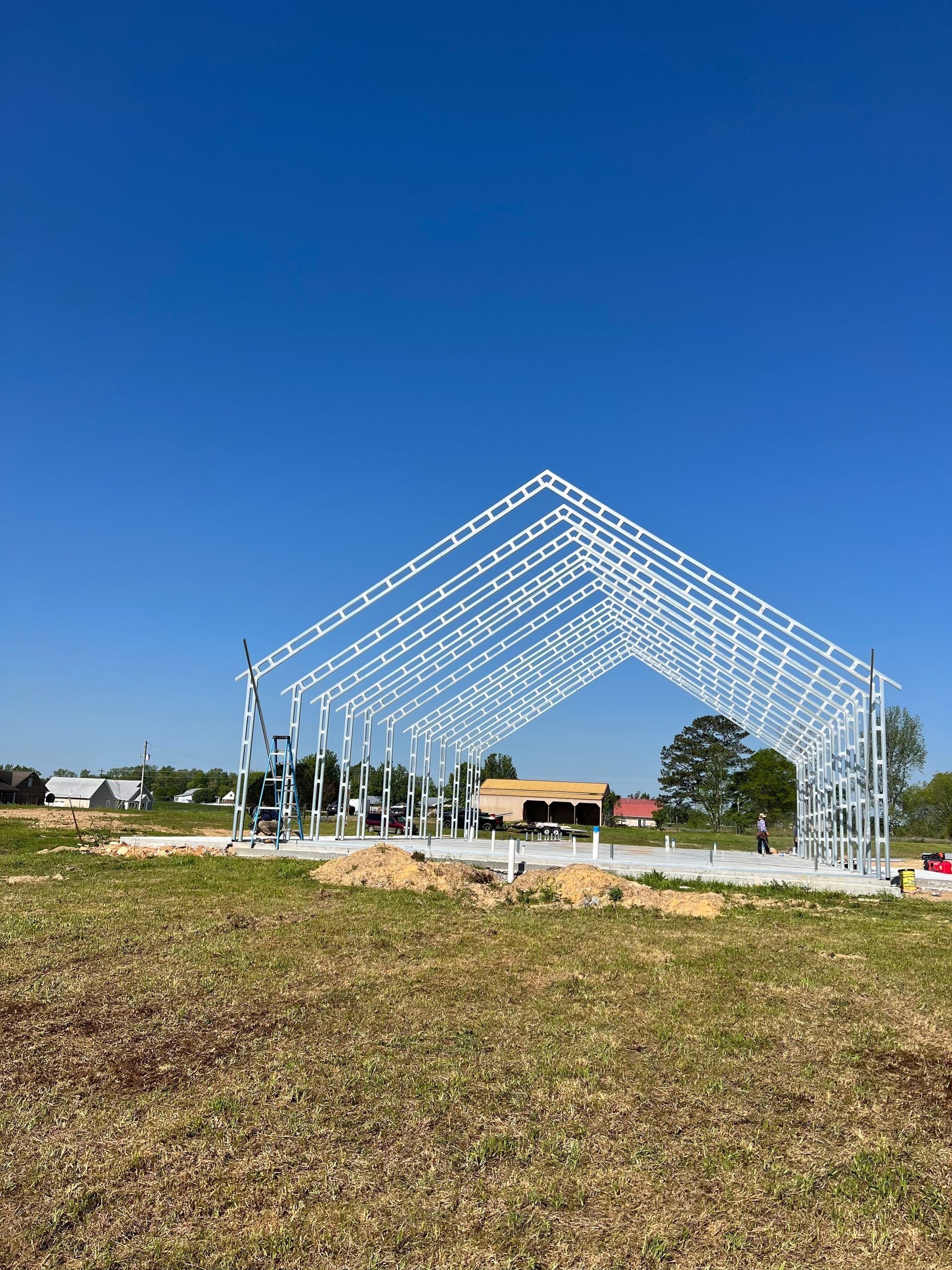 Steel framework of a building under construction on a concrete foundation, set in a grassy field under a blue sky.