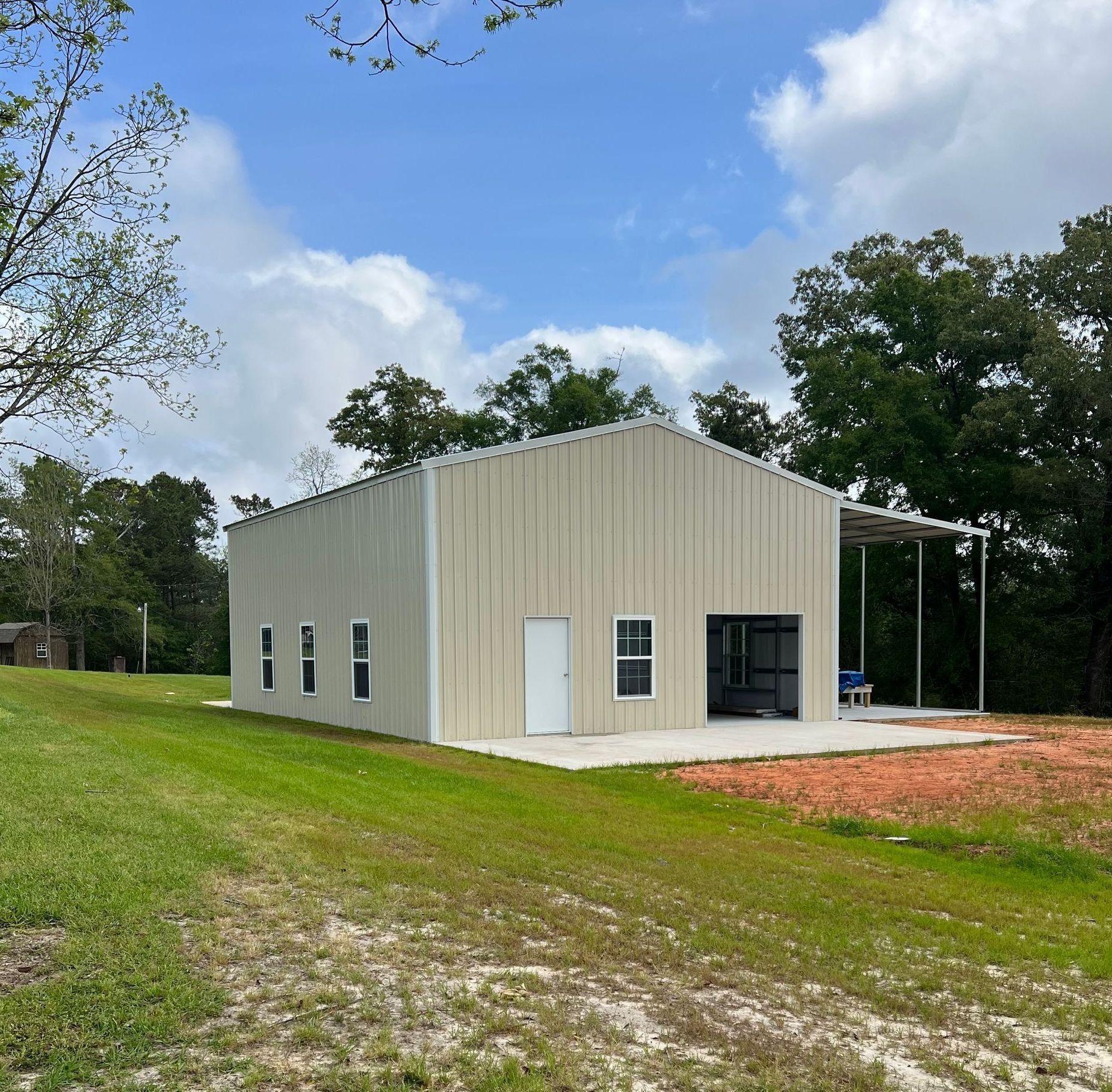 Beige metal building with open garage, small windows, and a porch on a grassy area under a blue sky.