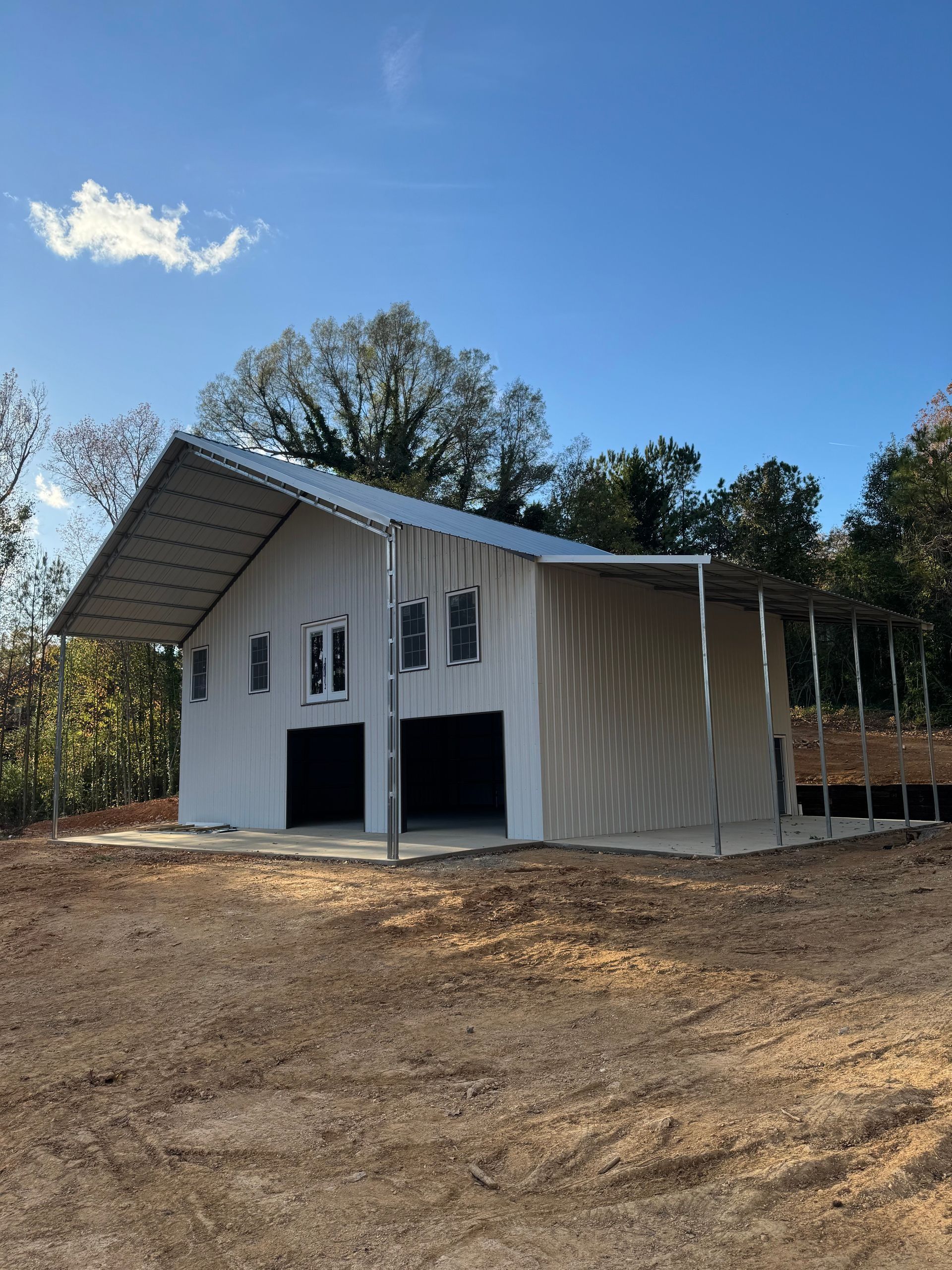 White barn with open bays, under construction, against a blue sky, surrounded by trees and dirt.