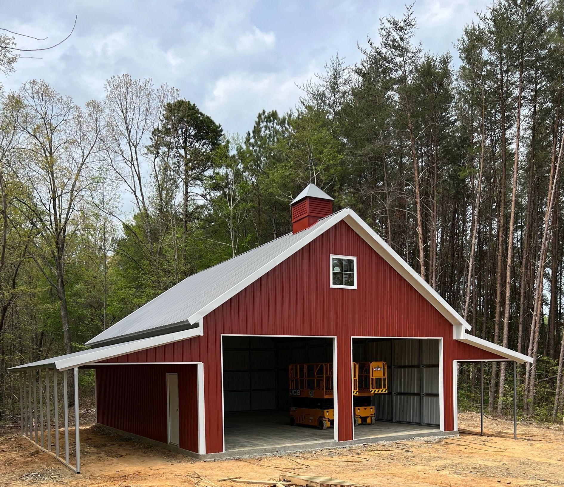 Red barn with two open bays, a cupola, and metal roof, set against a forest.