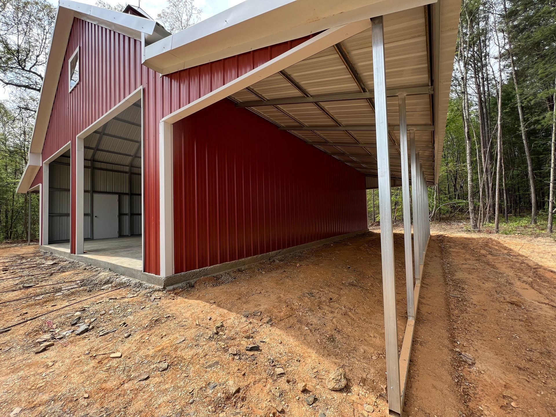 Red metal barn with attached carport; dirt ground, trees in background.