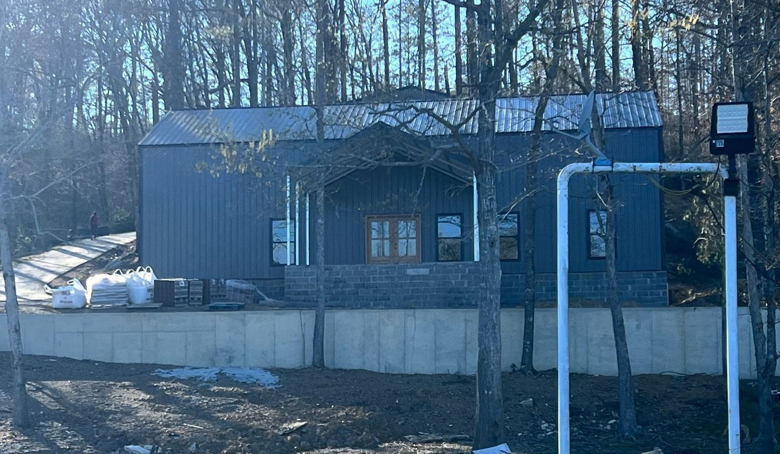 Blue-gray house with a stone porch, metal roof, and surrounding trees; under construction with materials visible.