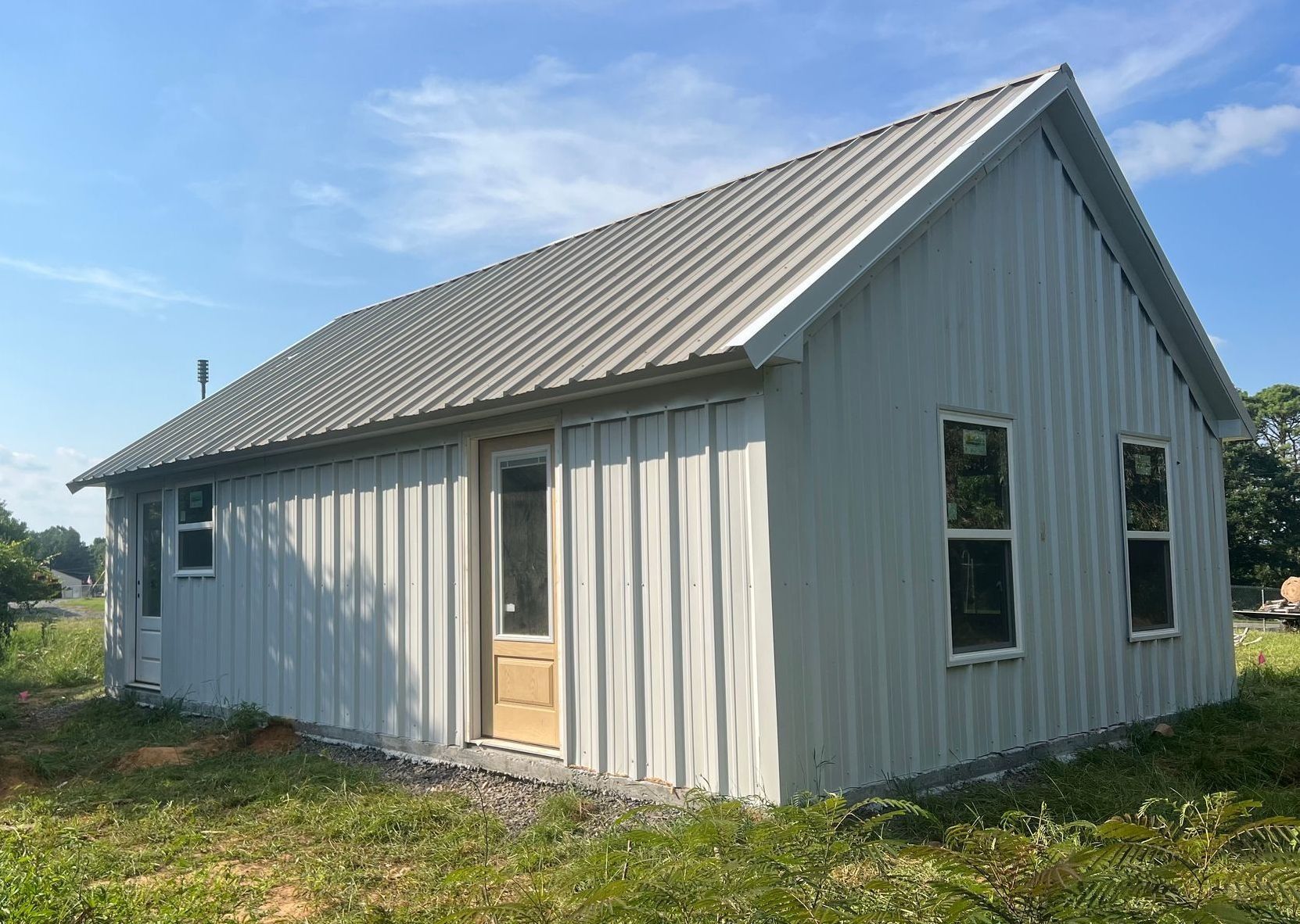 Gray metal-sided building with a gabled roof, two windows, and a doorway. Set in grassy area under a blue sky.